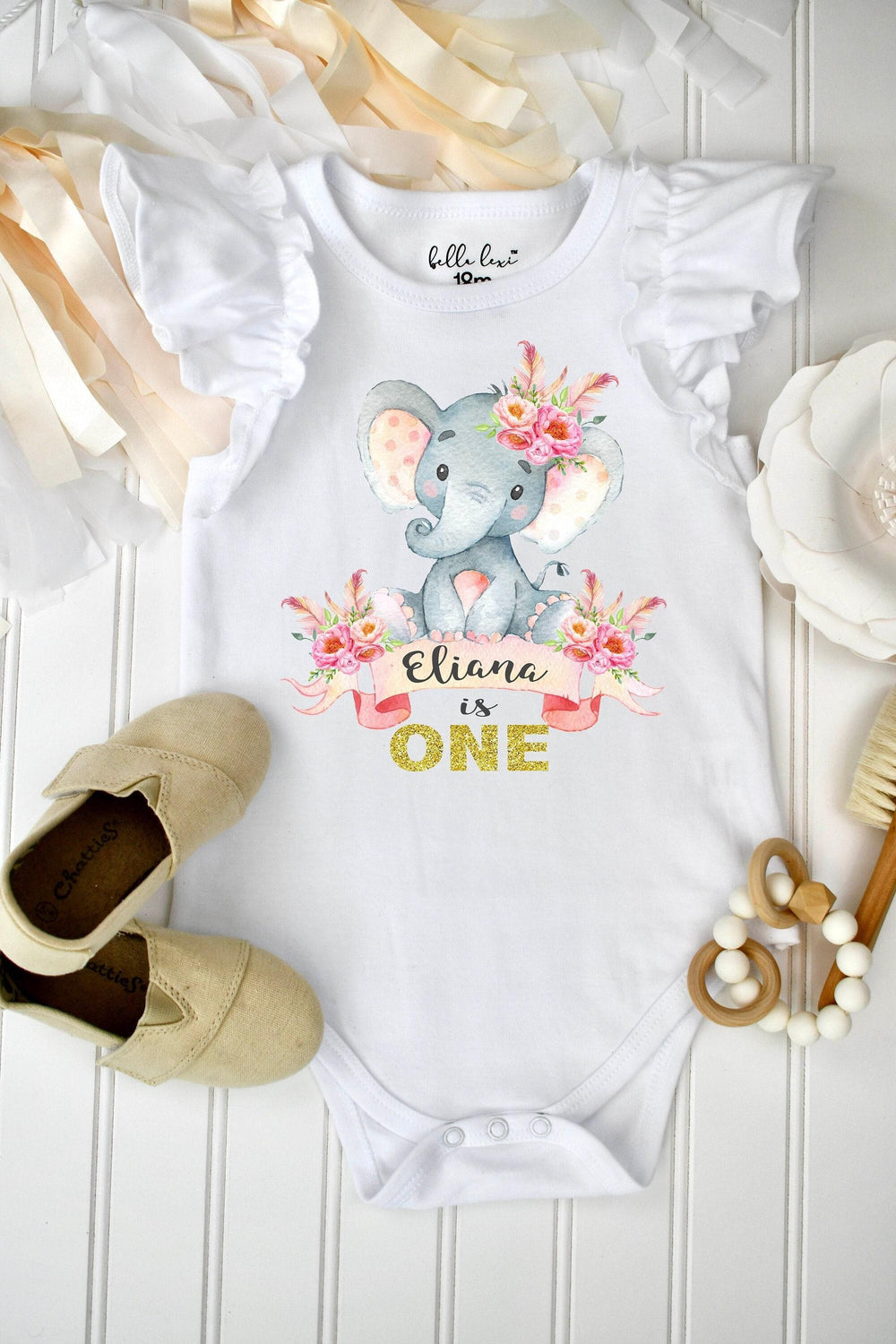 May include: A white short-sleeve bodysuit with ruffled shoulders featuring a graphic of a gray elephant with pink floral accents and feathers. The text reads Eliana is ONE in gold glitter lettering. The garment has a label reading Bella Lexi 18m.