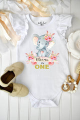 May include: A white short-sleeve bodysuit with ruffled shoulders featuring a graphic of a gray elephant with pink floral accents and feathers. The text reads Eliana is ONE in gold glitter lettering. The garment has a label reading Bella Lexi 18m.