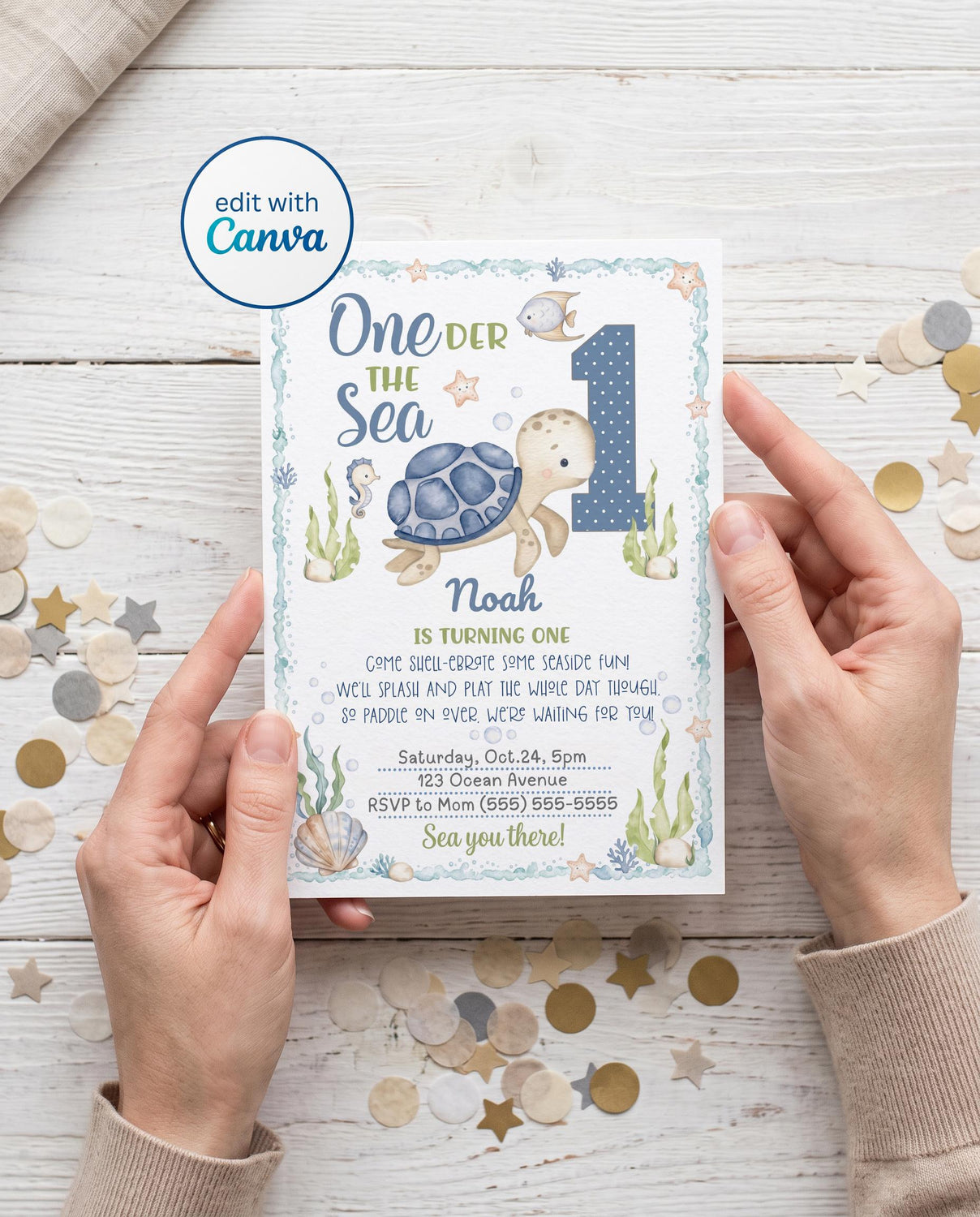 Hands holding a printed 5x7 One-der the Sea first birthday invitation with a watercolor sea turtle, blue polka dot number 1, seahorse, fish, starfish, seaweed, and seashells inside an ocean wave border, with an editable name, rhyming party wording, date, address, and RSVP, on a white wood table with confetti and an edit with Canva badge.