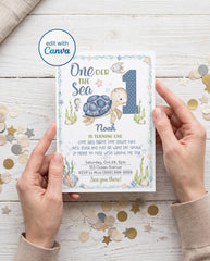 Hands holding a printed 5x7 One-der the Sea first birthday invitation with a watercolor sea turtle, blue polka dot number 1, seahorse, fish, starfish, seaweed, and seashells inside an ocean wave border, with an editable name, rhyming party wording, date, address, and RSVP, on a white wood table with confetti and an edit with Canva badge.