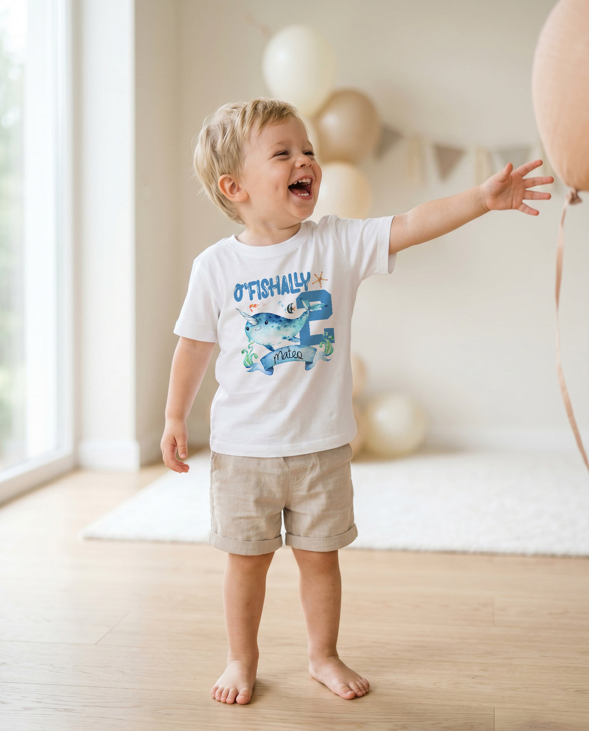 May include: A toddler wearing a white t-shirt with a blue narwhal graphic and the text "O'Fishally 2" and a blue and white party hat. The shirt has a white background with a blue narwhal and the text "O'Fishally 2" in blue. The toddler is standing in front of a blue and white background with white string lights.