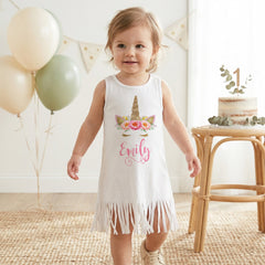 Child wearing a white dress with floral design and name 'Emily' in a room with balloons and a cake.